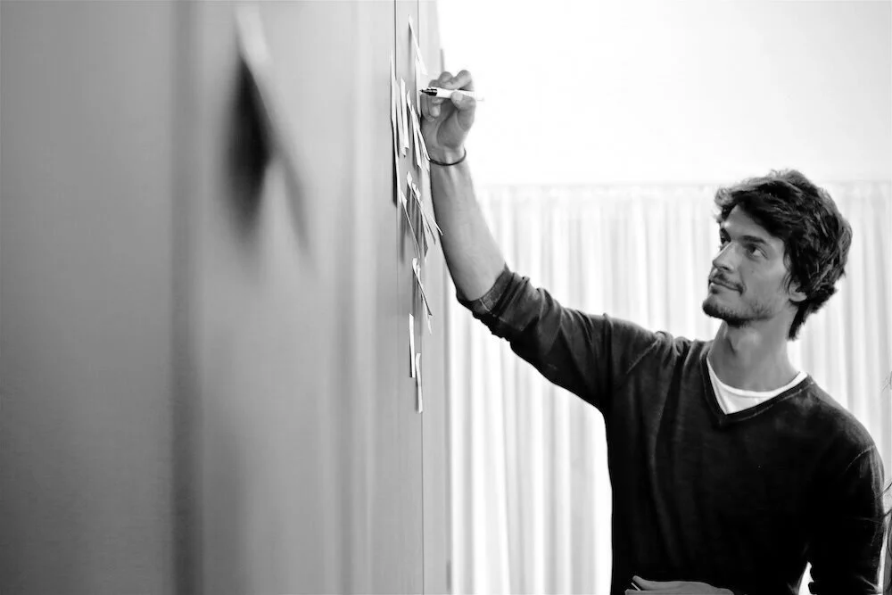 Schwarzweiß-Porträt von Korbinian Riedl an einem Whiteboard mit Klebezetteln, Co-Trainer der Evident Flow Ausbildung.
